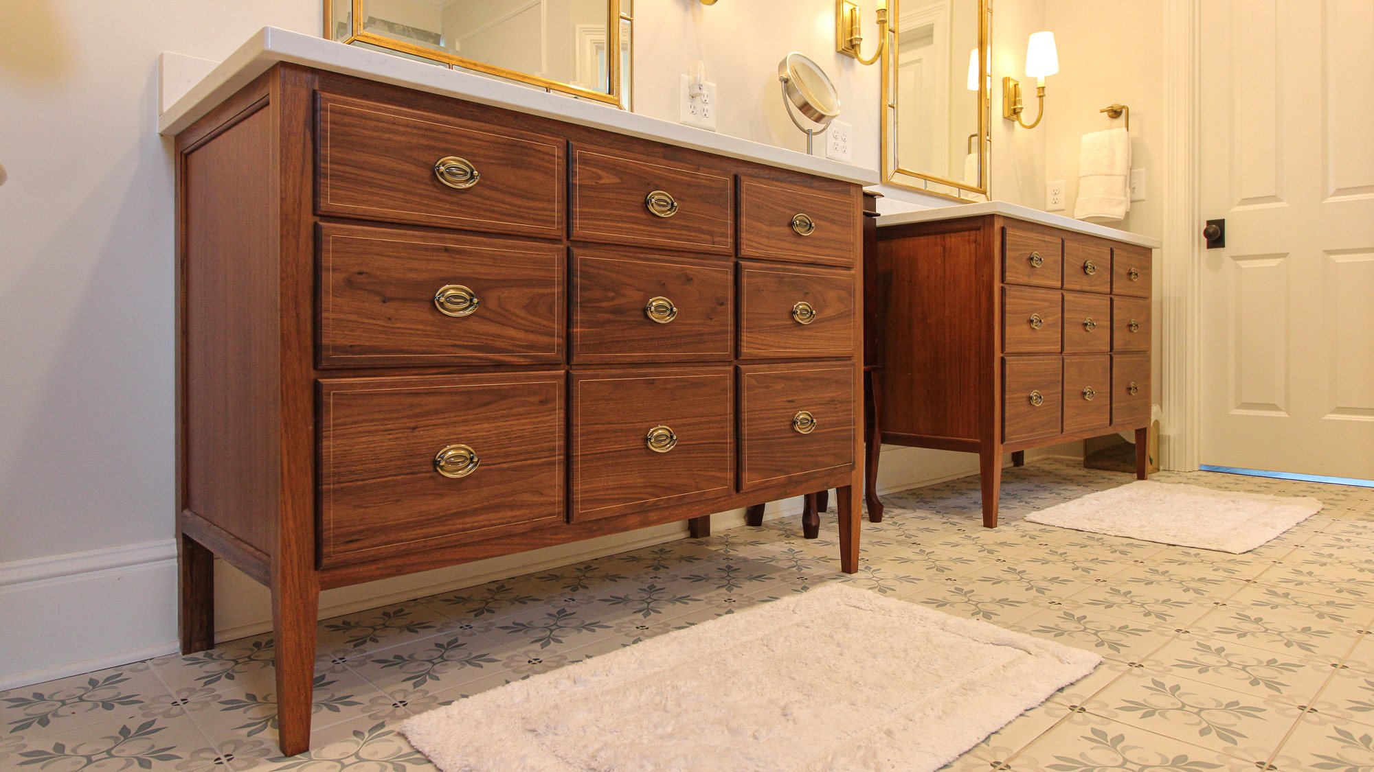 A pair of walnut bathroom vanities with inlaid string borders, oval brass pulls, tapered legs and white stone tops, installed in a tiled bathroom